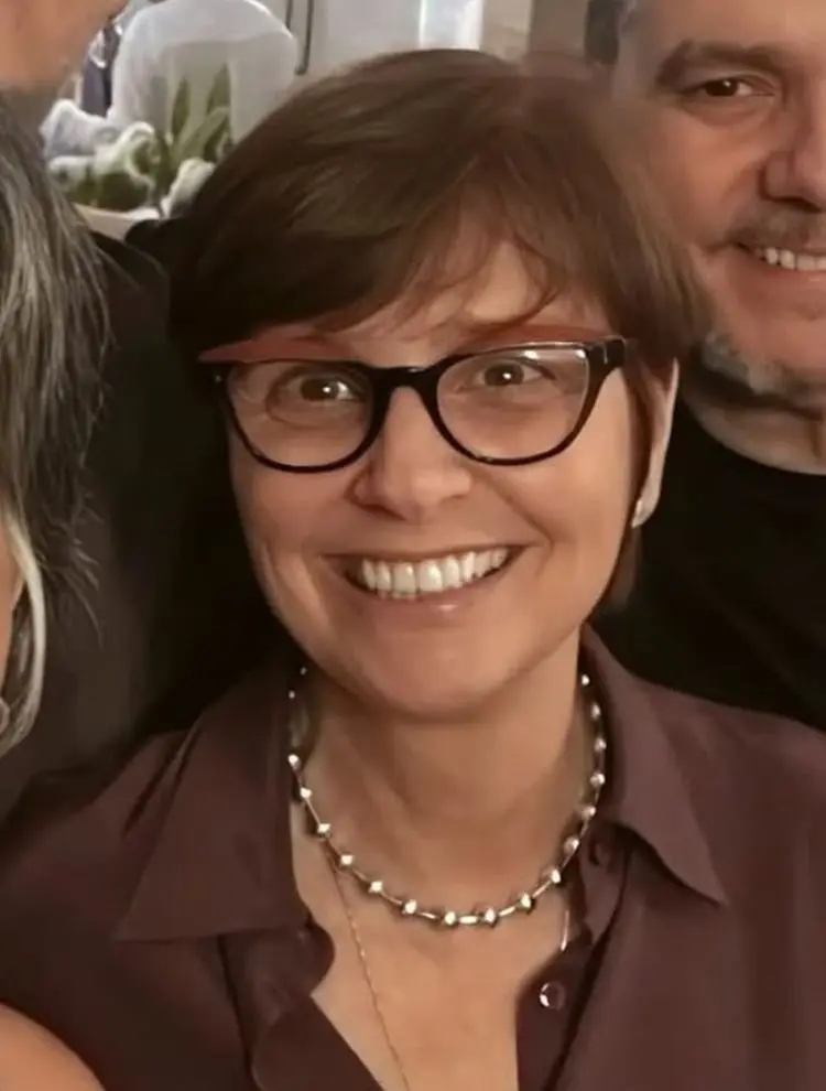 Mulher sorridente usa óculos de armação escura, cabelo curto castanho e colar de pérolas metálicas. Veste camisa marrom. Fundo desfocado mostra pessoas e plantas. Expressão alegre e amigável destaca-se como elemento principal na imagem.