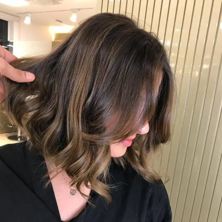 Uma mulher com um corte de cabelo bob ondulado e marrom com mechas sutis em tons mais claros, vista de perfil. Uma mão aparece no canto superior esquerdo ajustando o cabelo. Ela usa uma blusa escura.
