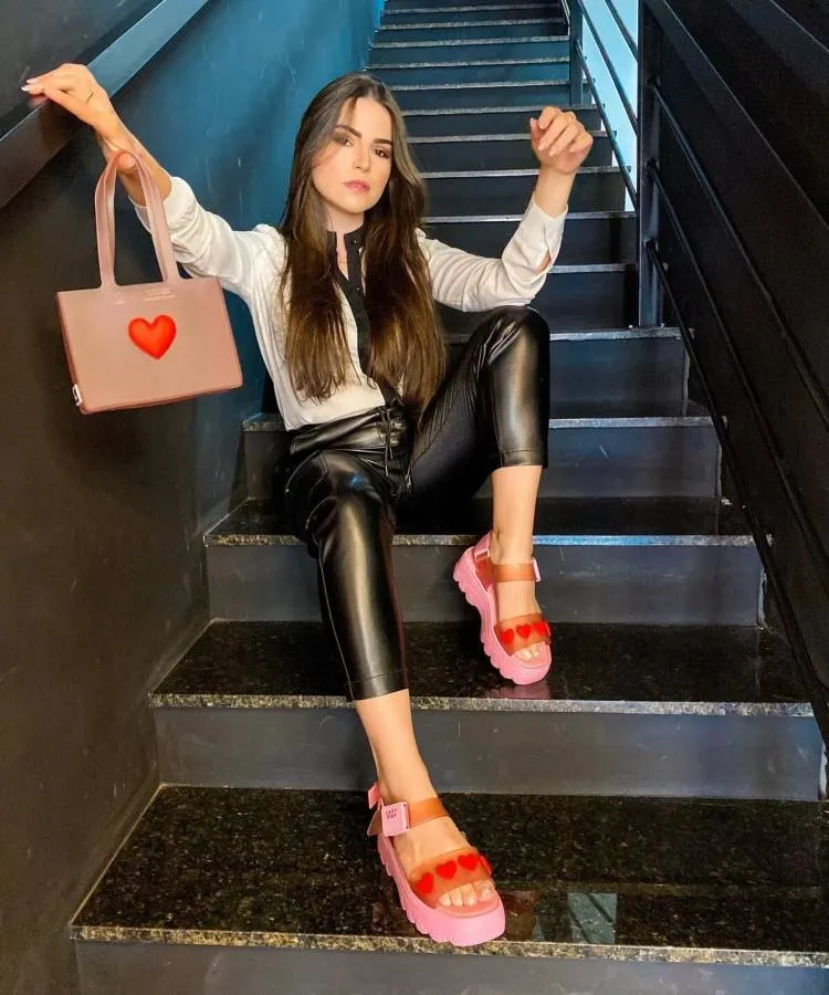 Uma mulher branca de cabelos longos posa sentada em uma escada, ela usa uma calça preta, uma blusa branca e papetes rosa