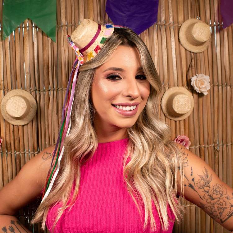 Uma mulher loira sorridente, com maquiagem marcante e tatuagens nos braços, veste um top rosa vibrante. Ela usa um mini chapéu de palha de festa junina decorado com fitas coloridas. Ao fundo, uma parede de bambu decorada com bandeirinhas juninas e pequenos chapéus de palha.
