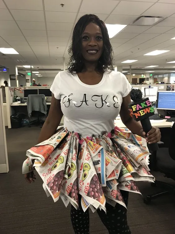 Mulher de pele negra com blusa branca escrito "Fake", saia de jornal e microfone escrito "fake news"