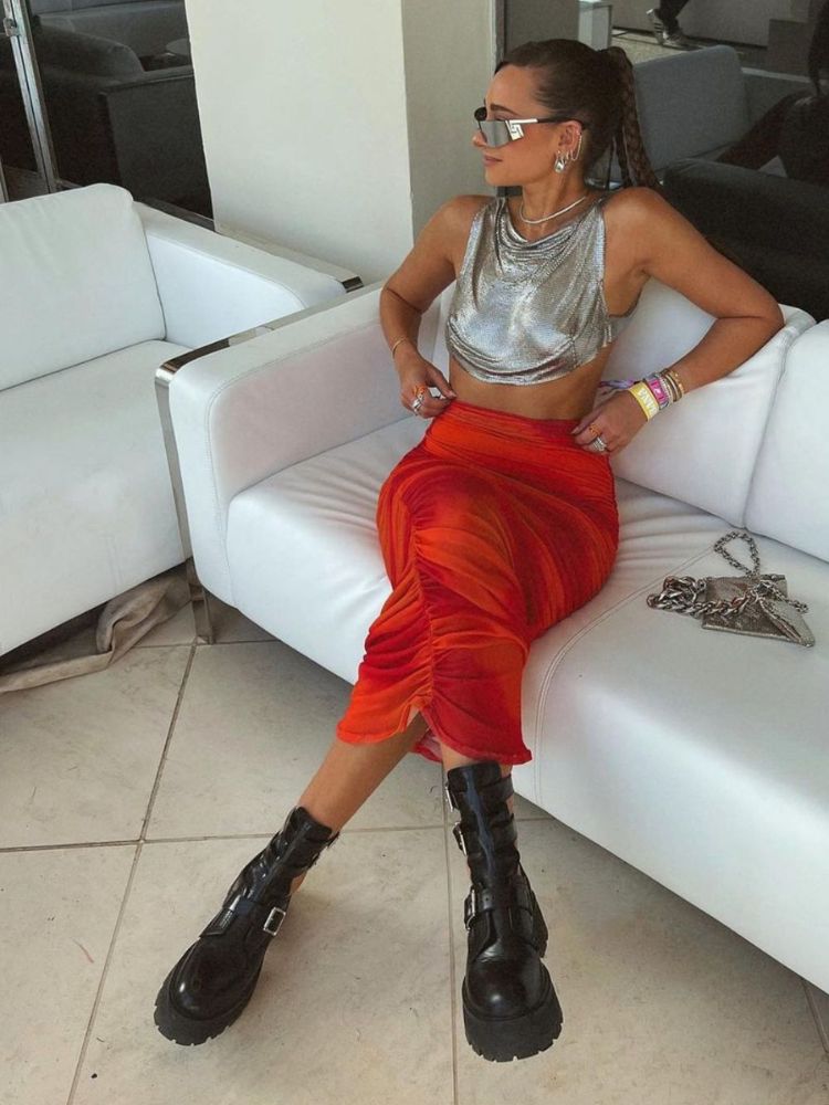 Mulher de pele clara usando top metalizado e saia midi laranja com bota preta