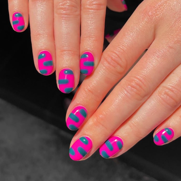 Mãos exibindo estilos de unhas curtas, com design moderno em esmalte rosa vibrante e padrões geométricos ondulados em azul-petróleo. O acabamento brilhante destaca o contraste das cores. Fundo preto desfocado, enfatizando o visual ousado e criativo das unhas.