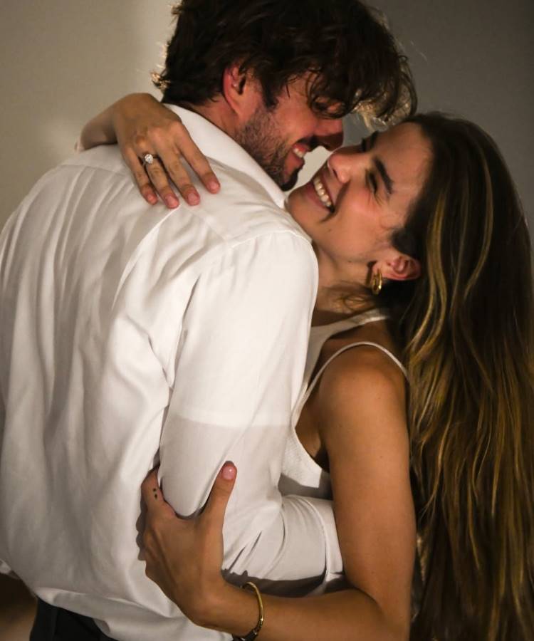 Um casal feliz se abraça, com ambos sorrindo e se olhando. A mulher usa um anel de noivado com uma pedra brilhante em sua mão esquerda, enquanto o homem a segura. O clima é de intimidade. Famosas que estão noivas