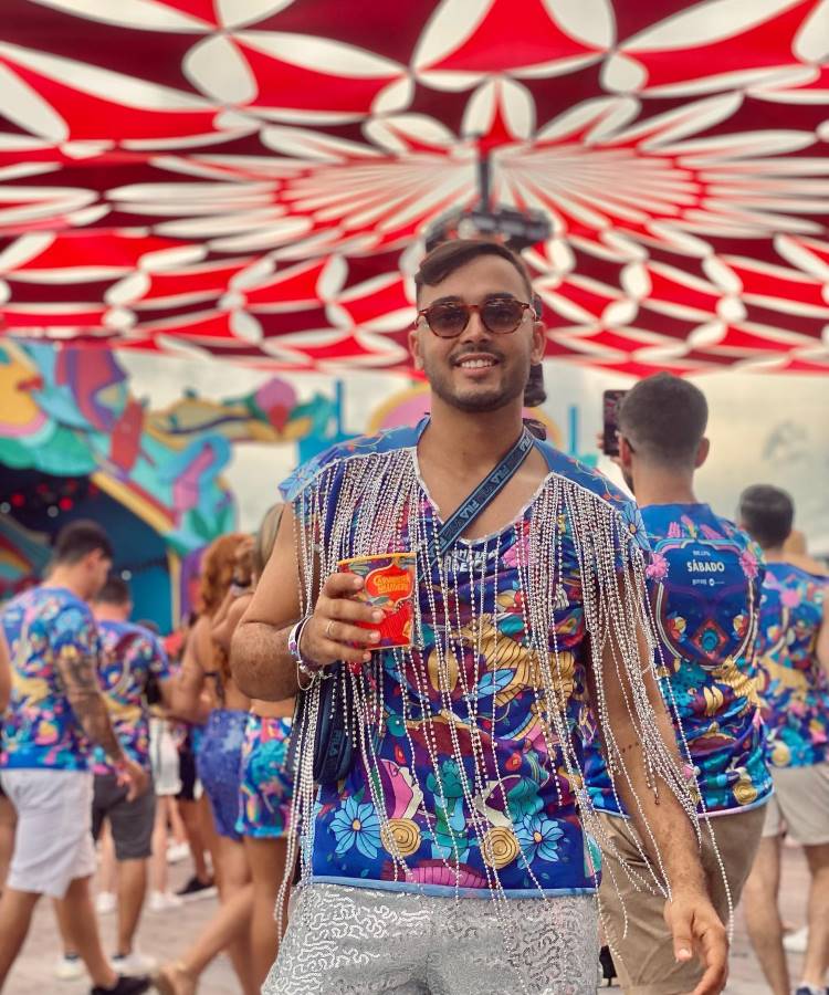 A imagem mostra um homem sorridente em um ambiente de festa, vestindo um abadá vibrante decorado com franjas brilhantes. Ele segura uma bebida em lata, com um colorido cenário carnavalesco ao fundo e um toldo estampado acima.
