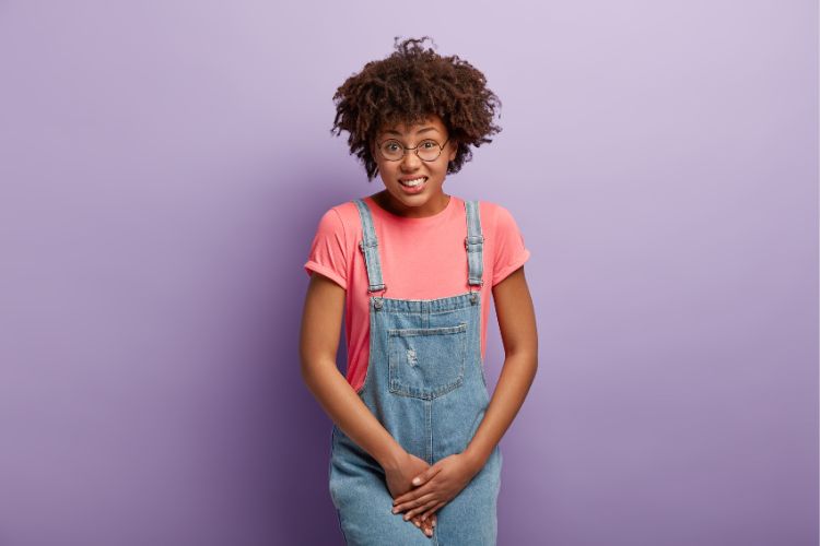 Mulher de pele negra usando blusa rosa e jardineira jeans longa, com as mãos na região íntima e semblante de sofrimento.