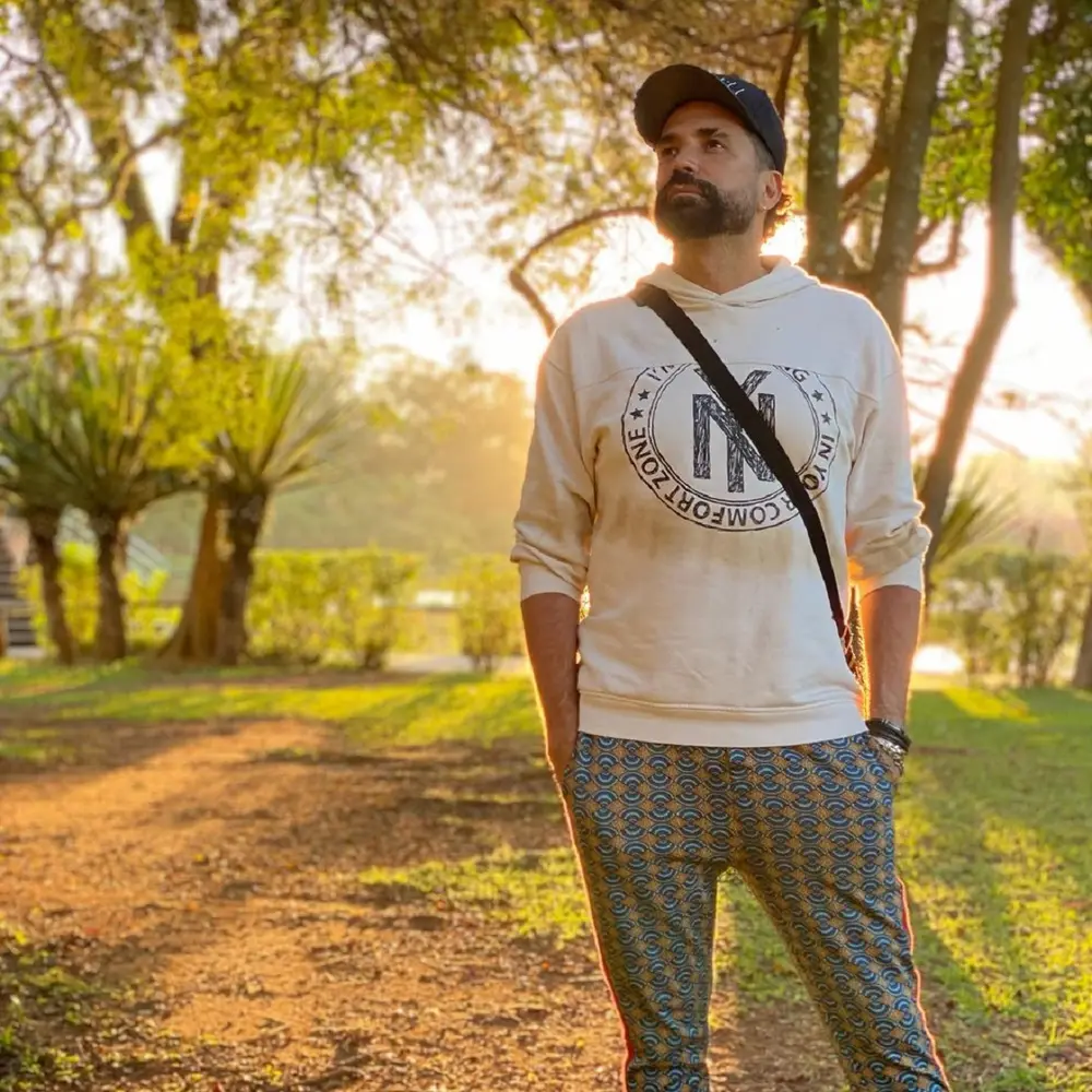 Foto de Latino com expressão séria, de boné, moletom, calça estampada e bolsa tiracolo. De fundo, tem-se a imagem de um parque ou jardim