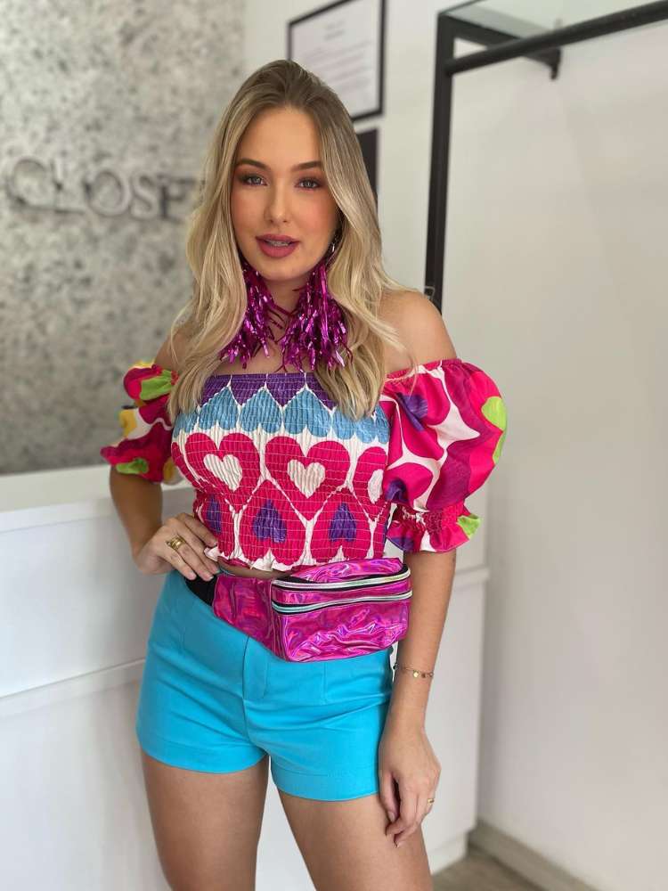 Mulher de pele clara e cabelo loiro usando shorts azul, cropped de corações coloridos, pochete roxa e brinco de fitas roxas. 