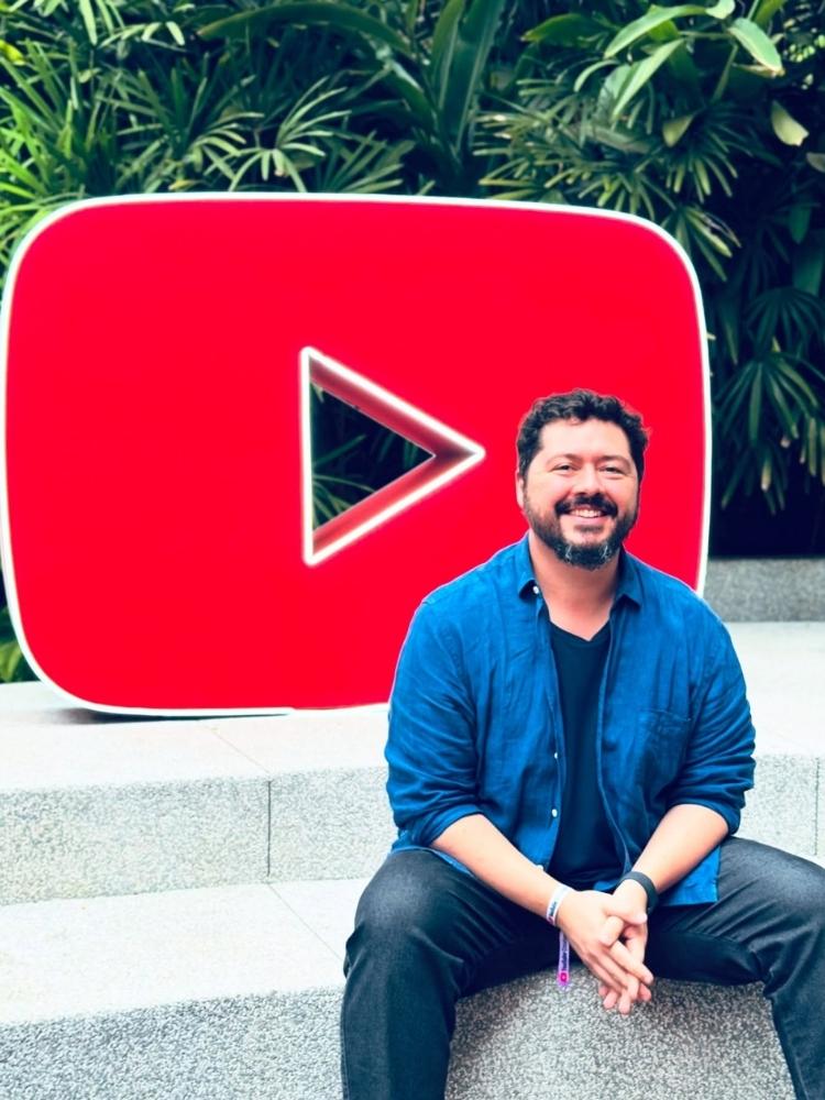 Atila usando jaqueta azul, camiseta escura, calça jeans, sentado em escada branca com logo do YouTube grande atrás