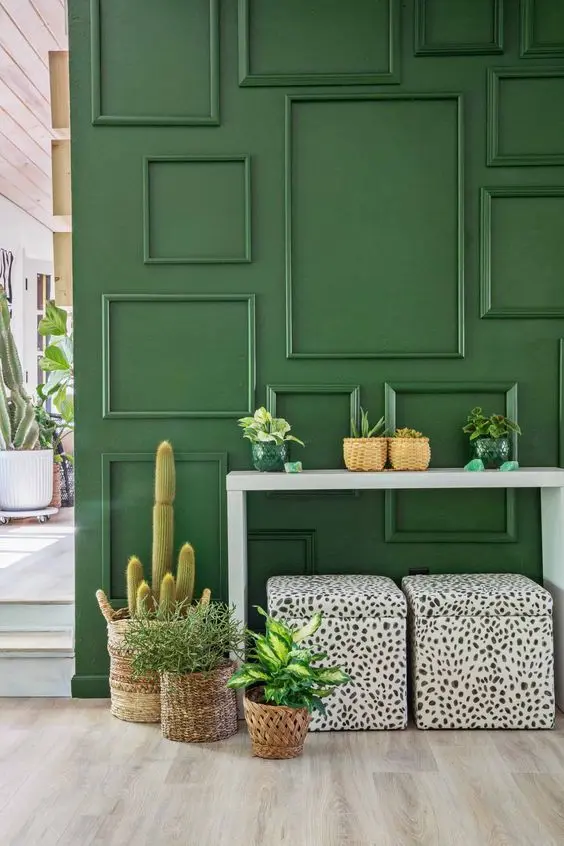 Hall com parede verde com moldura boiserie, aparador branco, plantas e puffes de onça