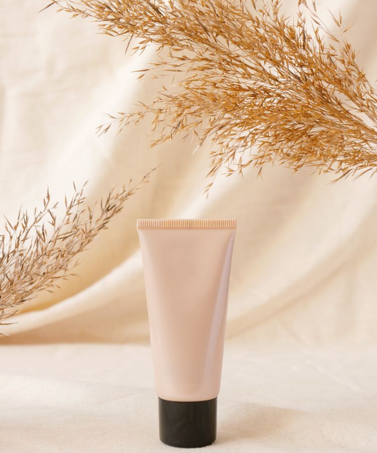 A imagem mostra um tubo de base em creme com tampa preta, posicionado verticalmente sobre uma superfície clara. Ao fundo, há ramos secos que emolduram o produto. Este é um dos tipos de base que proporciona cobertura suave e é ideal para um acabamento natural na maquiagem.


