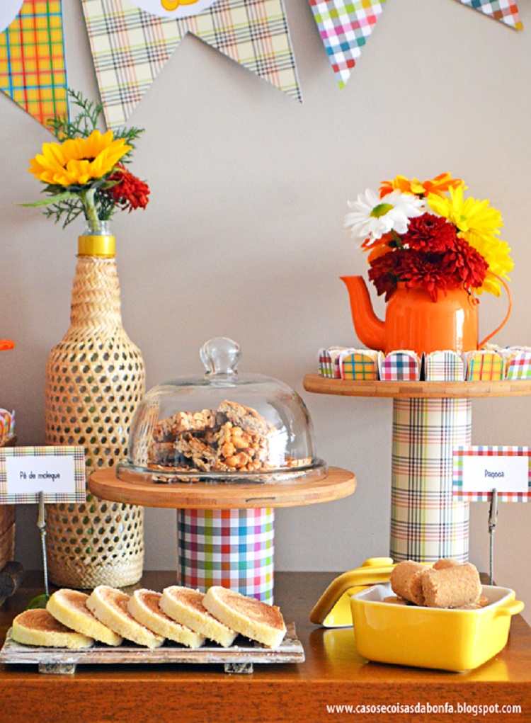 A imagem representa festa junina decorada com flores. Contém mesa com bolos, paçoca e pé de moleque