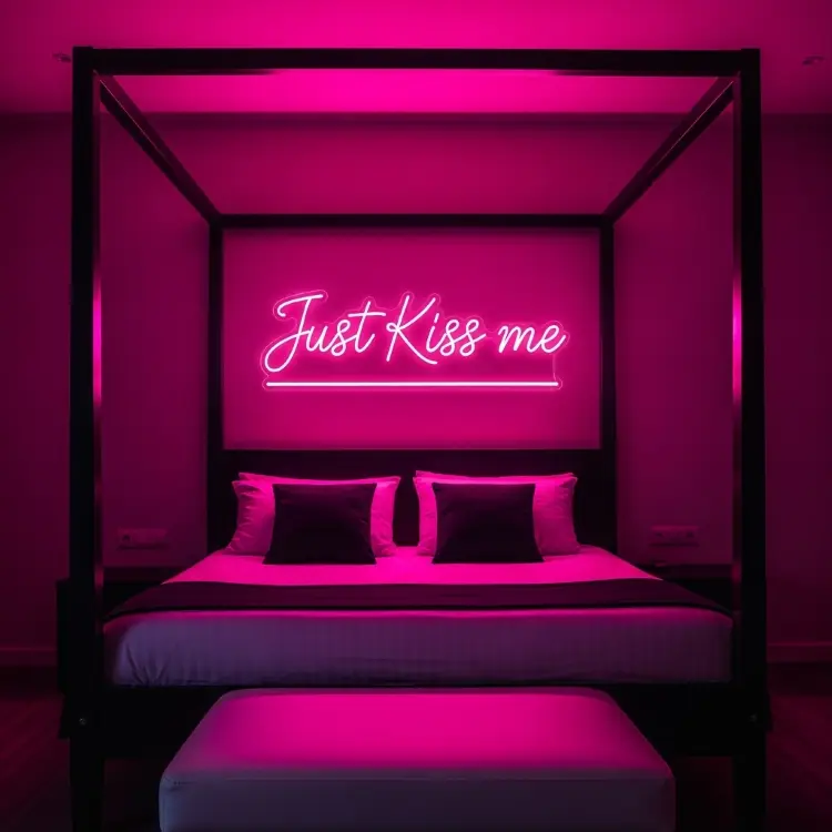 Uma cama de dossel preta. Na cabeceira, a parede é iluminada por um letreiro de neon rosa que diz "Just Kiss me". A cama tem lençóis brancos e travesseiros escuros. Um banco estofado está aos pés da cama.
