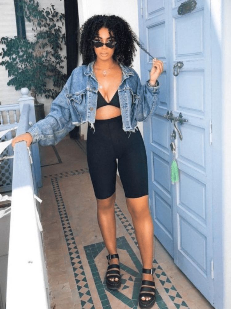 Mulher de pele negra com bermuda ciclista, top e jaqueta jeans cropped. 