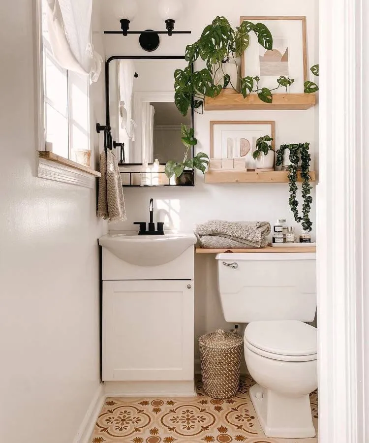 Um banheiro aconchegante com piso estampado em tons de bege e marrom. Possui um armário branco com pia, um espelho retangular preto, prateleiras de madeira com plantas e quadros. Um cesto de vime e toalhas dobradas complementam o ambiente.

