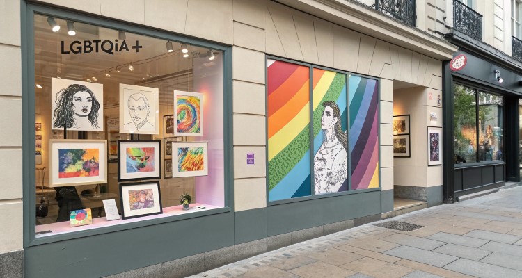Fachada de loja LGBTQIA+ com duas vitrines: uma exibe ilustrações variadas, incluindo retratos e arte abstrata; a outra apresenta um painel com arco-íris vibrante e a ilustração de uma pessoa estilizada. Calçada urbana ao redor. Ambiente acolhedor.

