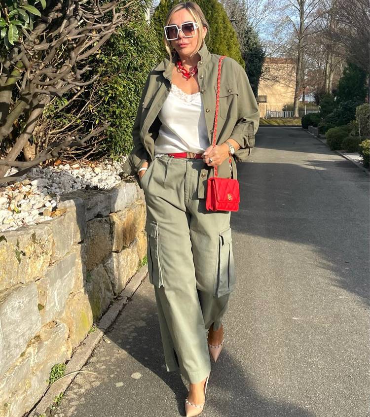 Mulher madura usando regata branca, calça cargo verde, camisa verde com cinto vermelho, bolsa vermelha e scarpin, andando em rua pública.