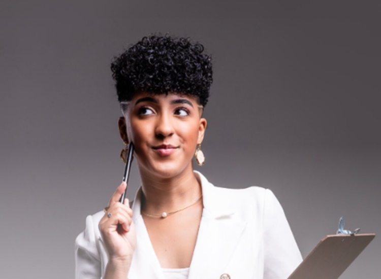 A imagem mostra Nath Finanças sorrindo, com cabelo curto e encaracolado. Ela usa um blazer branco, segura uma prancheta e está com uma caneta perto do rosto, transmitindo uma expressão pensativa. O fundo é cinza neutro.


