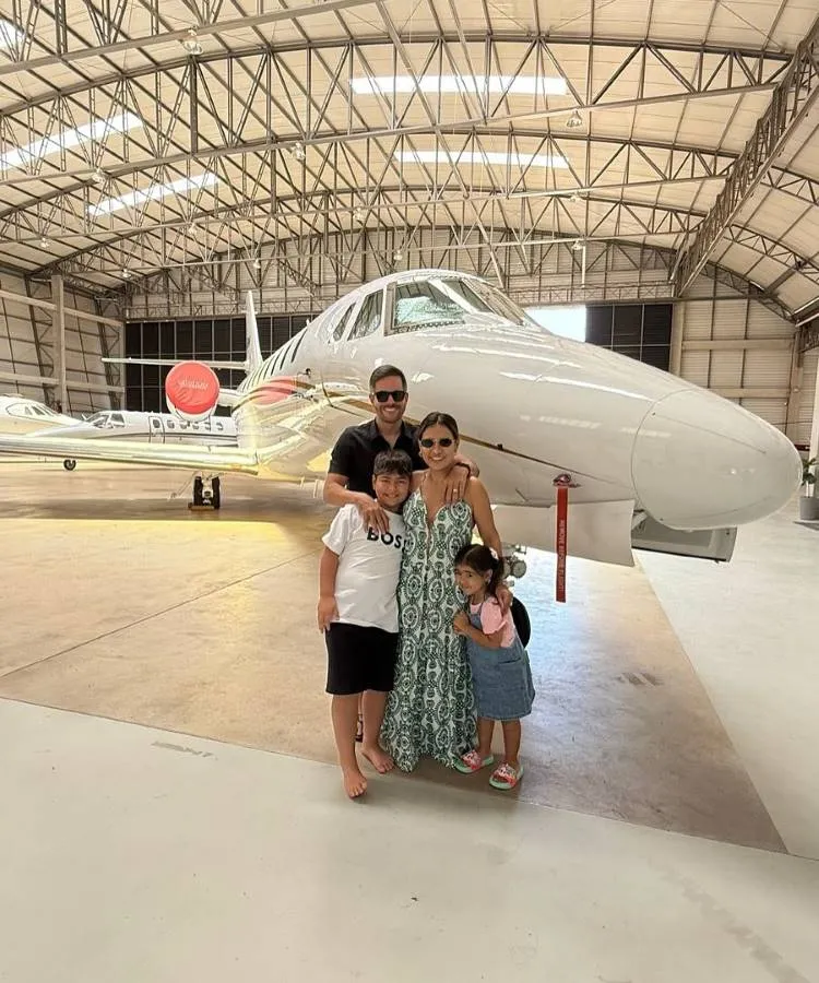 Uma família feliz de quatro pessoas, um pai, uma mãe e dois filhos, posa na frente de um avião particular branco dentro de um hangar. Todos usam óculos de sol e sorriem para a câmera, com o pai abraçando a mãe.


