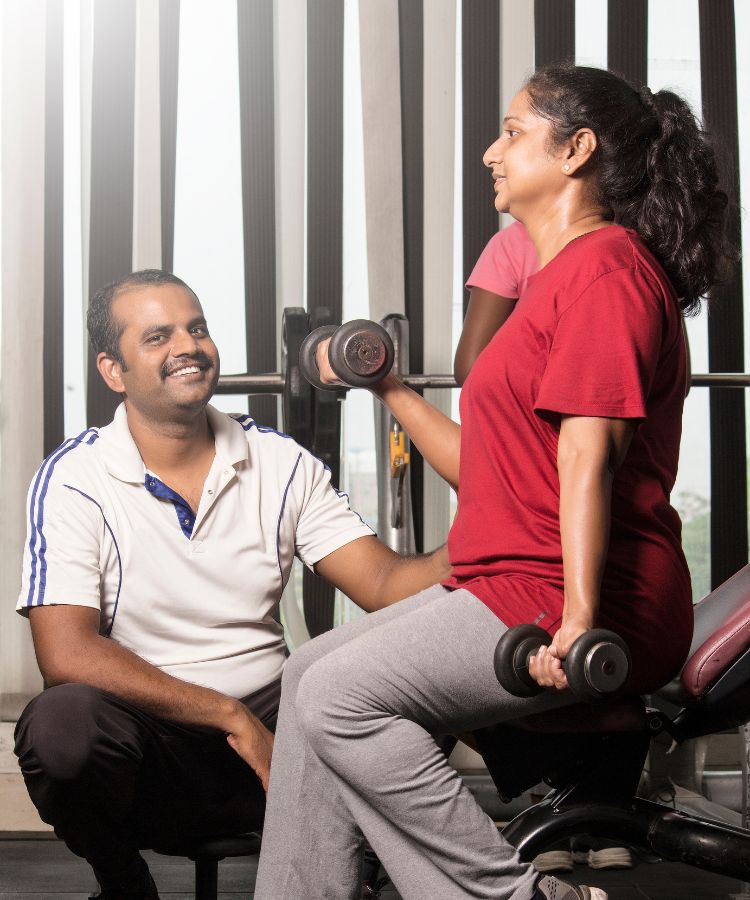 A imagem mostra duas pessoas em um ambiente de academia. Uma pessoa está sentada em um banco de exercícios, segurando halteres e parece estar se exercitando. A outra pessoa está ao lado, aparentemente auxiliando ou orientando o exercício.