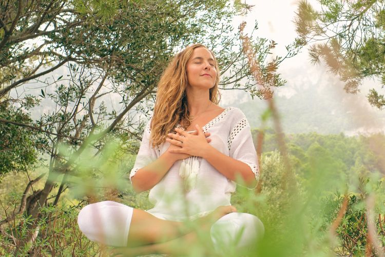 Mulher sentada na natureza, em posição meditativa, com as mãos sobre o coração e semblante sereno. Cercada por árvores e luz suave, está conectada à energia espiritual, representando sintonia e equilíbrio durante a abertura do portal 3/3.





