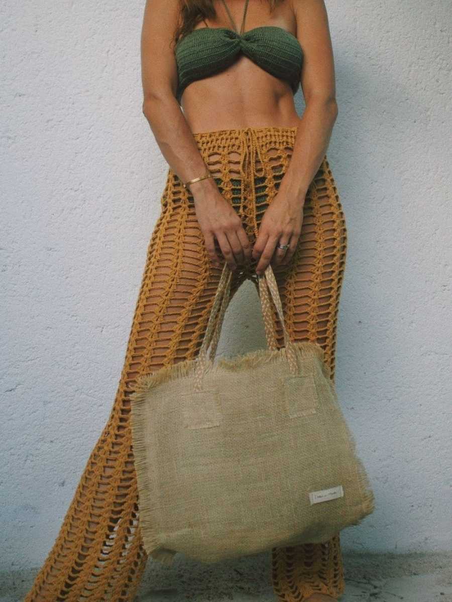 Mulher fotografada do pescoço aos pés, usando biquíni verde de crochê e saída de praia amarela de crochê. Além disso, segurando bolsa artesanal.
