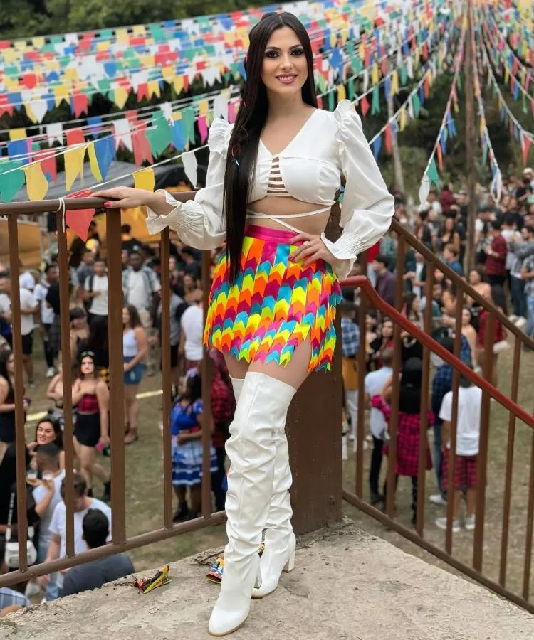 A imagem mostra uma festa junina animada. Uma mulher, de sorriso aberto, veste blusa branca e saia colorida, com botas brancas. Ao fundo, bandeirinhas coloridas decoram o local movimentado, cheio de pessoas em trajes típicos, refletindo alegria e tradição cultural.
