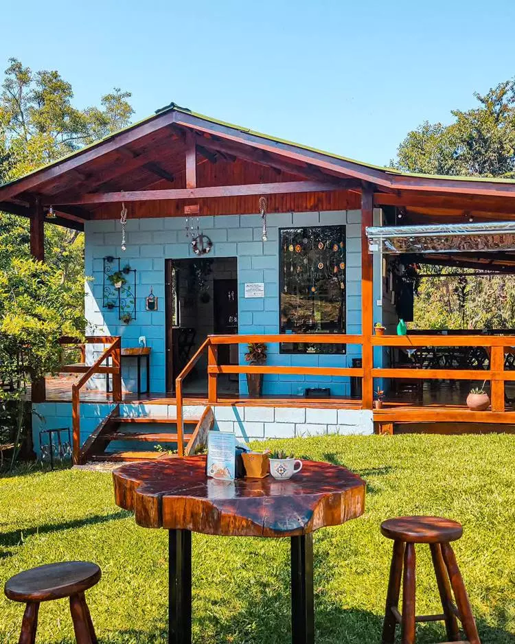 Imagem de fachada de casa simples azul com escada e detalhes em madeira envernizada. No jardim gramado, uma mesa de madeira com dois bancos também de madeira envernizados. Sob a mesa, xícara com planta e outras decorações.
