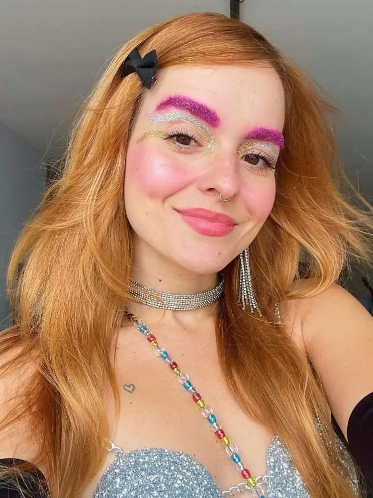Mulher de pele clara usando sombra roxa e glitter na sobrancelha além de delineado gráfico grosso prateado e dourado