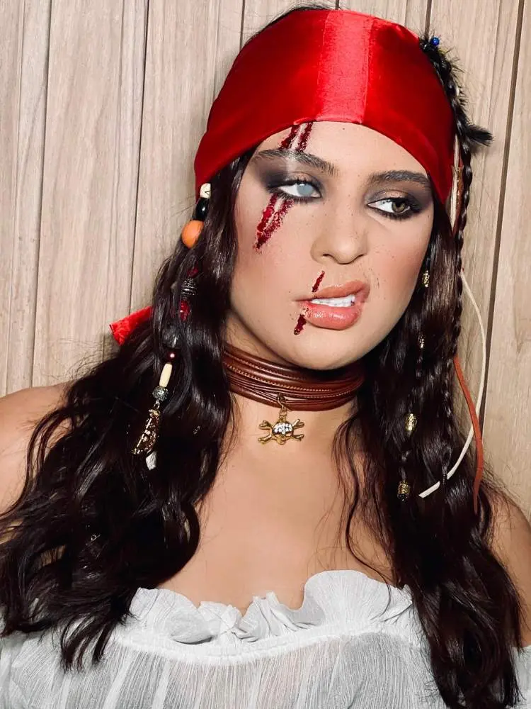 Mulher de pele clara fantasiada de pirata, com lenço vermelho, lente imitando olho de vidro, machucado com sangue falso no olho e na boca