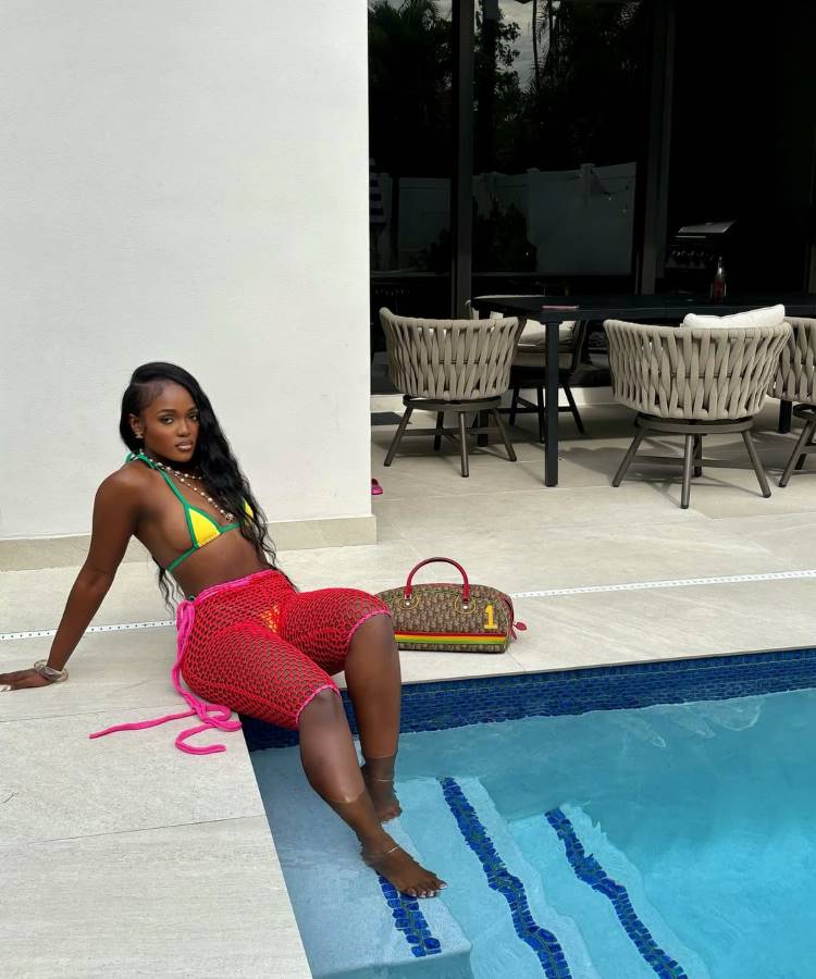 Mulher sentada na beira de uma piscina, com um pé na água. Ela usa biquíni e uma saída de praia de crochê vermelha. Ao lado dela, uma bolsa. Ao fundo, cadeiras e uma mesa.