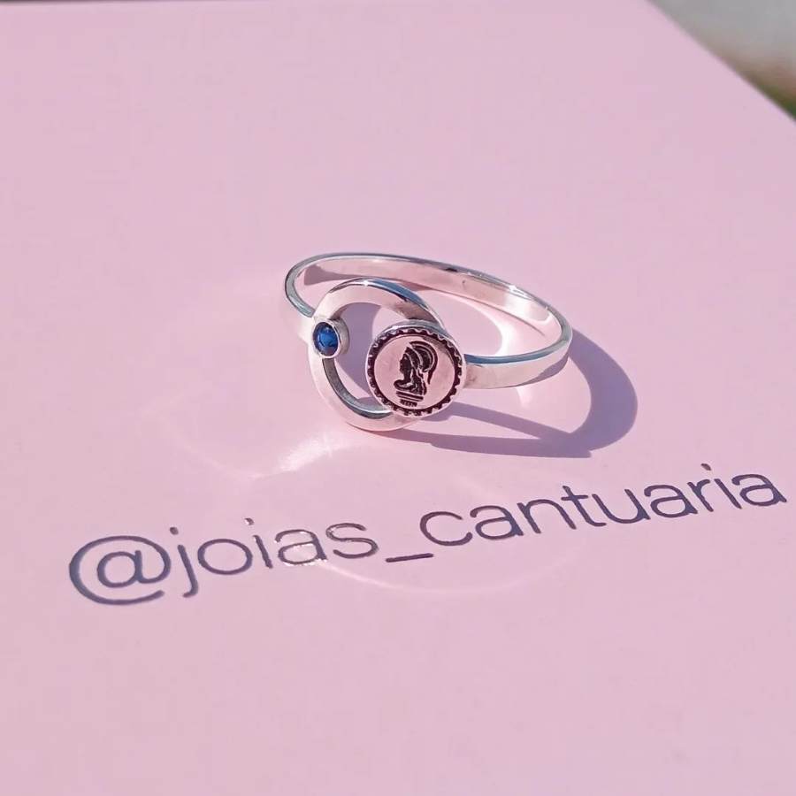Anel fino prateado com pequena pedra azul e símbolo de engenharia