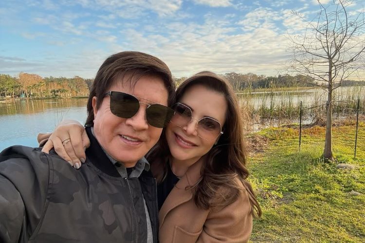 Xororó e Noely SORRI para uma selfie em frente a uma paisagem natural com lago, árvores e céu parcialmente nublado. Ambos usam óculos escuros; ele veste jaqueta preta, e ela, casaco bege. Cena transmite tranquilidade.