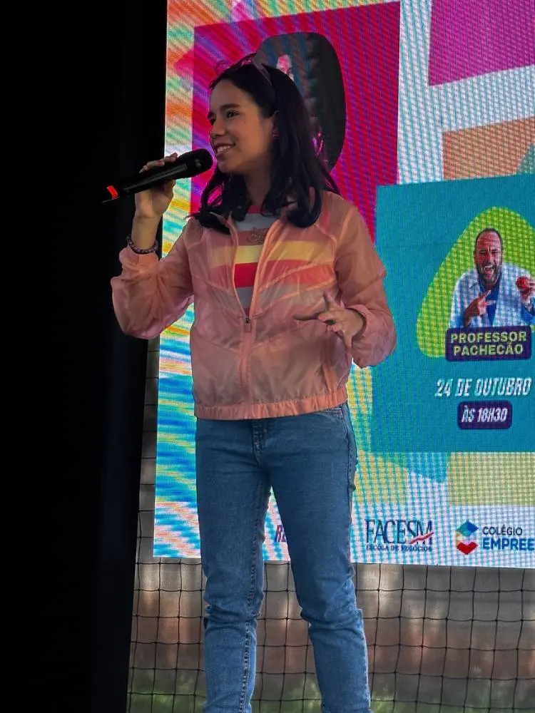 Malu Lira de blusa colorida, casaco rosa, calça jeans, em palco palestrando 