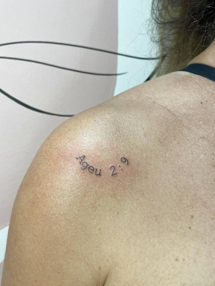 Mulher de pele clara com tatuagem escrita fine line "Ageu 2:9"