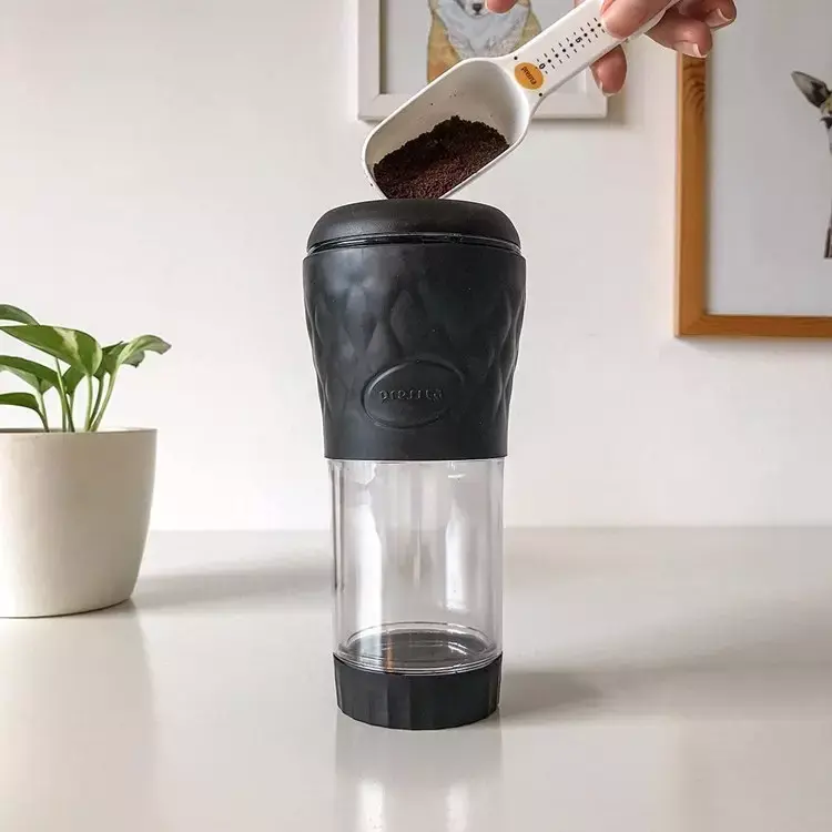 Cafeteira portátil sob superfície com vaso e planta. Há uma mão adicionando pó de café com colher medidora