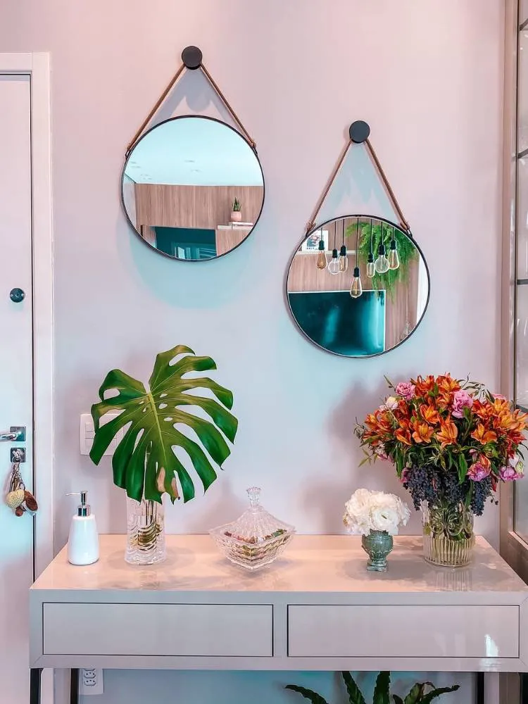Aparador com dispenser branco, vaso transparente com costela-de-adão, baleiro quadrado, outros dois vasos enfeitados e dois espelhos redondos pendurados. 