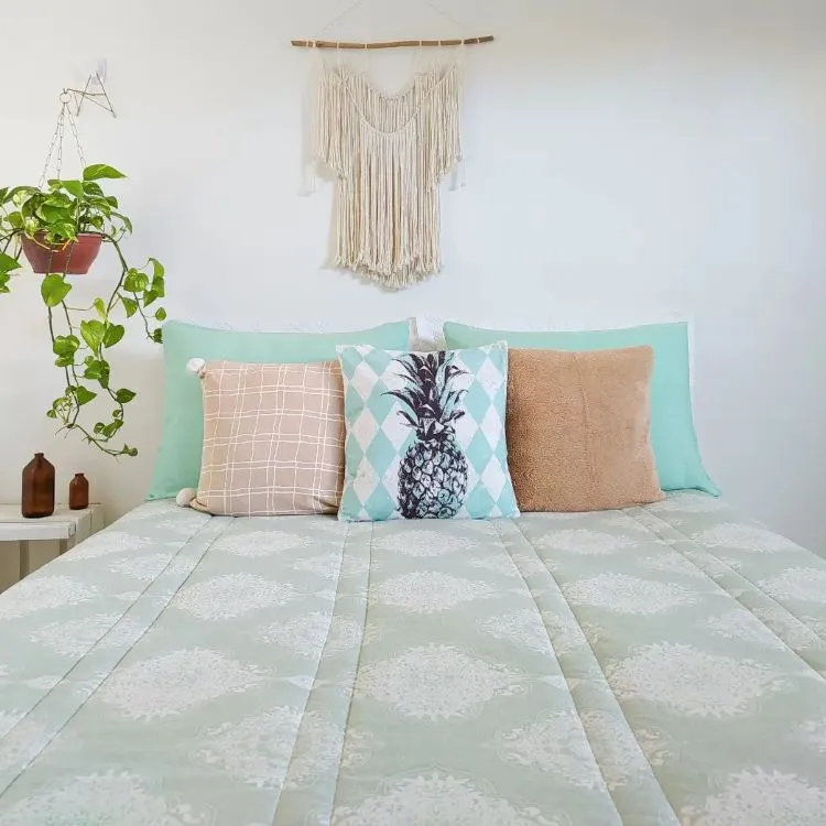 A imagem mostra uma decoração fácil para quarto com uma cama de frente para a câmera. A cama tem uma colcha geométrica verde e branca, três almofadas (uma com estampa de abacaxi), um enfeite de parede em macramê e um vaso suspenso com planta verde.

