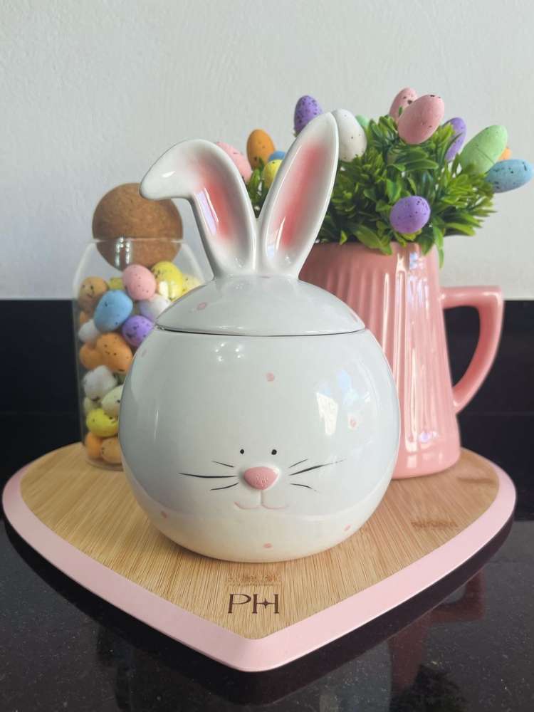 Tábua de madeira em coração com pote branco de coleho, jarra rosa com arranjo de plantas e ovos coloridos e pote de vidro com ovos coloridos.