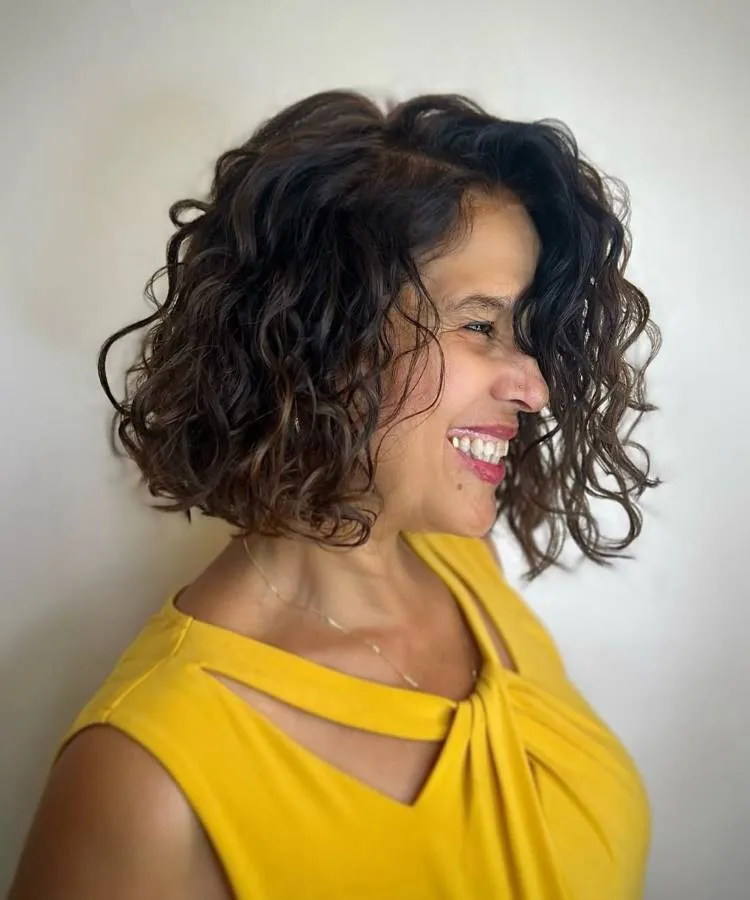 Uma mulher sorridente com um corte de cabelo cacheado, na altura dos ombros, posa de perfil. Ela tem cachos castanhos escuros e veste uma blusa amarela vibrante com recortes no decote.