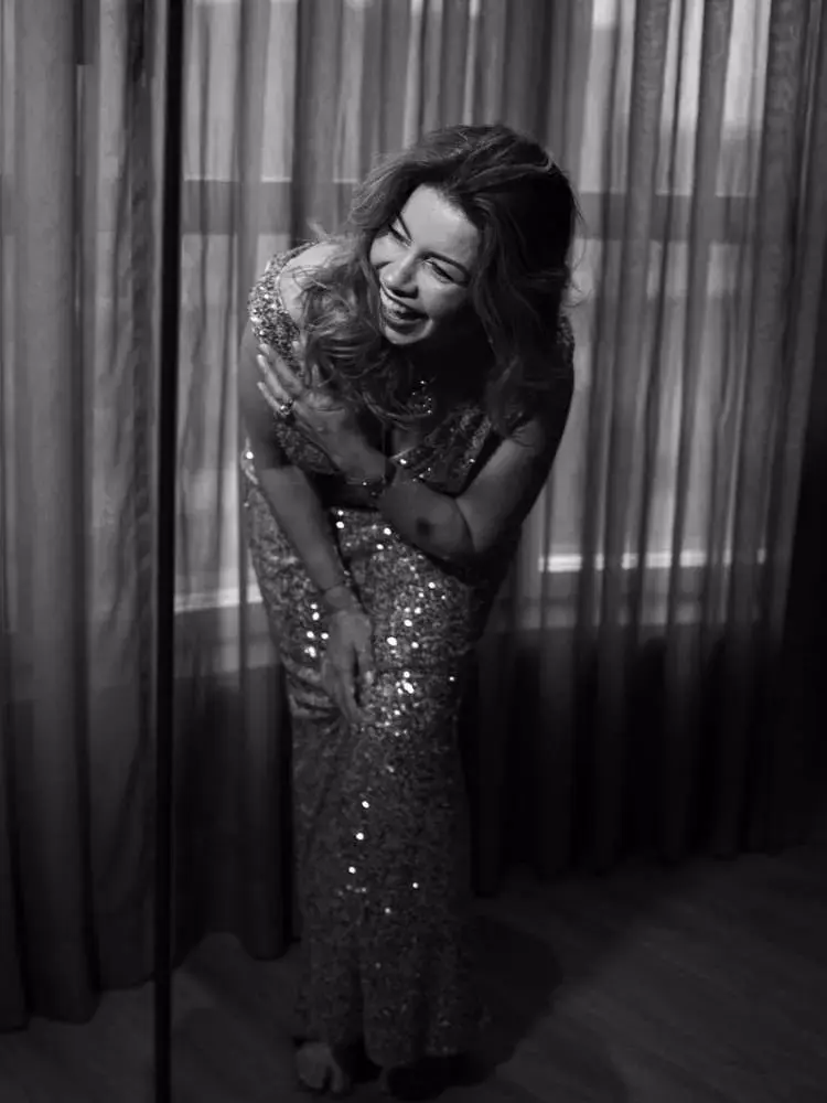 Mulher não-modelo com mais de 40 anos em ensaio fotográfico, usando vestido de paetês dourado, em banheiro. Foto preto e branco. Ela sorri naturalmente