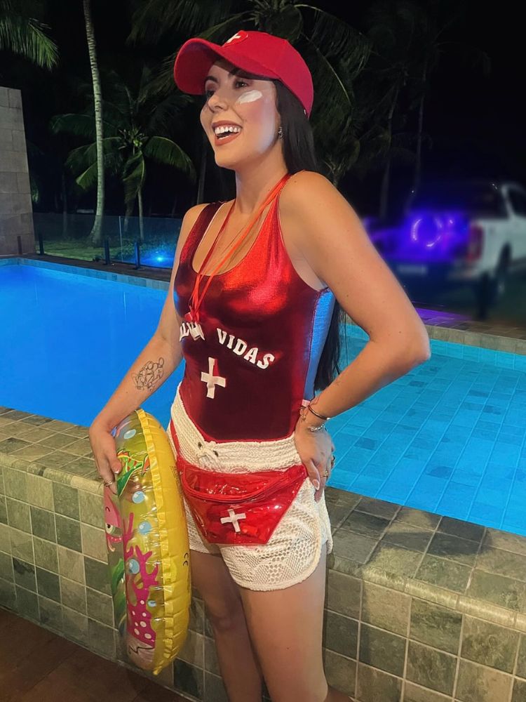 Mulher de pele clara com body vermelho escrito salva-vidas com cruz branca, boné vermelho, shorts branco, apito, pochete vermelha e boia