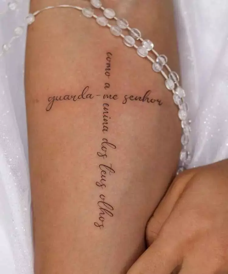 A imagem mostra uma tatuagem elegante em forma de cruz com a frase: "guarda-me Senhor como a menina dos teus olhos". A escrita cursiva é suave e delicada. O braço está adornado com um bracelete de contas transparentes, complementando o visual.
