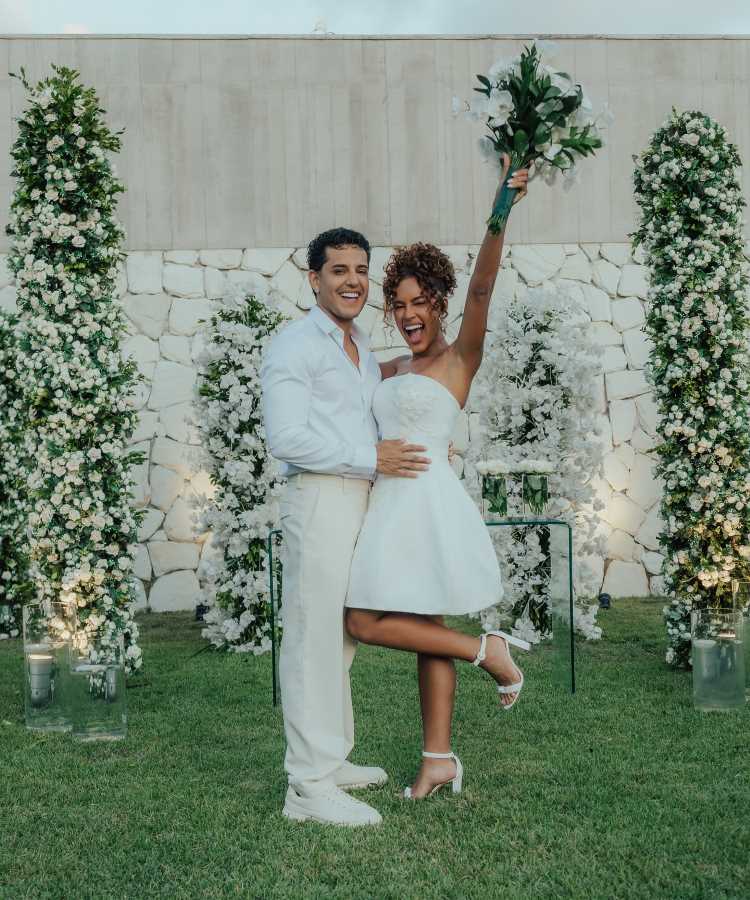 Casal Sthe Matos e Kevi Jonny celebra união em casamento deslumbrante ao ar livre, com decoração exuberante em branco e verde. Noiva veste vestido curto, segura buquê e sorri animada. Noivo em traje claro. Imagem ilustra os casamentos de famosos 2025.
