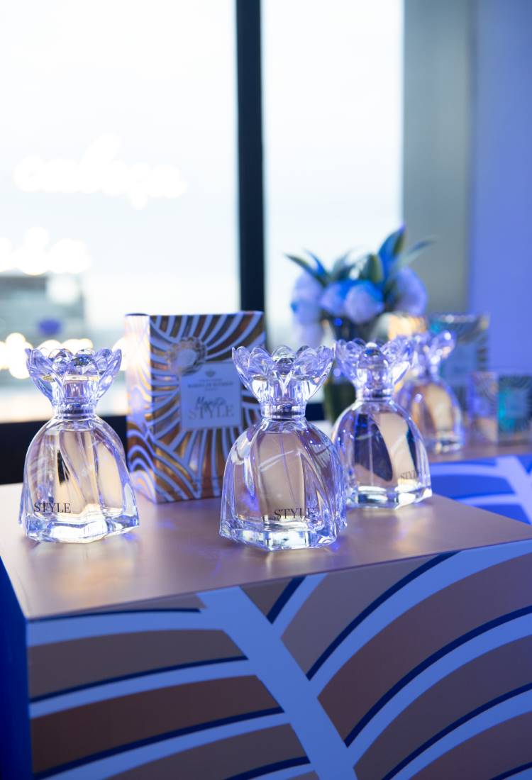 Frascos de perfume cristalinos com tampa decorativa em uma mesa coberta por padrão azul e dourado. Ao fundo, embalagem do perfume e flores em um vaso. A iluminação azul cria uma atmosfera elegante e sofisticada, destacando o produto.
