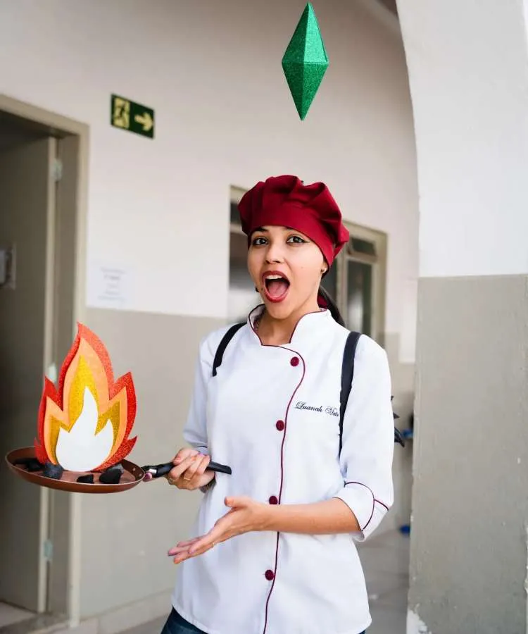Fantasia, cosplay de mulher chef do The Sims, para o Halloween, usando tiara de prisma verde, vestindo doma branca e segurando frigideira que simula fogo de E.V.A