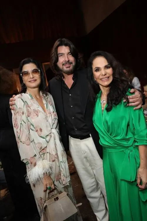 Tania Derani de óculos com vestido floral de plumas e segurando bolsa, Stephane Rolland de terno preto e calça off-white e Cris Porto de verde em fundo escuro