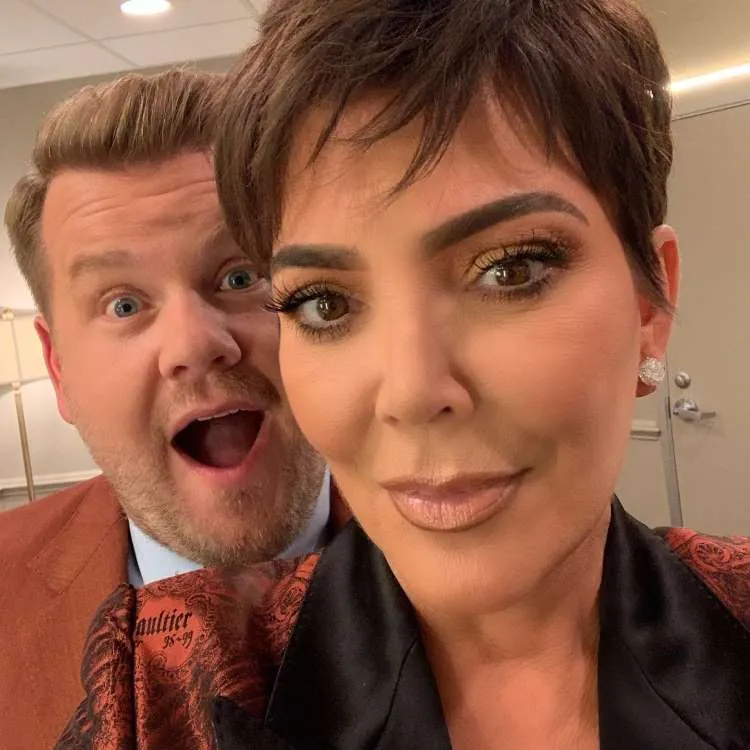 Kris Jenner tira uma selfie com James Corden, ambos com expressões de surpresa. Ela usa uma maquiagem dourada nos olhos e um blazer estampado, exibindo seu corte de cabelo pixie.