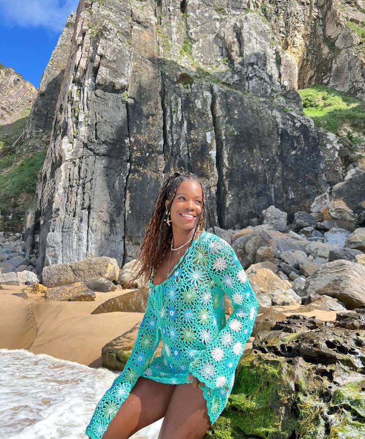Uma mulher negra, com tranças no cabelo, sorri para a câmera. Ela usa uma saída de praia de crochê verde-água e branco com padrão de flores, sentada em uma rocha na praia com falésias rochosas ao fundo.