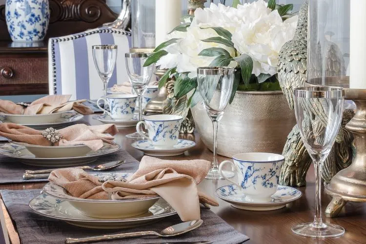 A imagem mostra uma mesa de madeira. Há quatro pratos brancos com o mesmo padrão floral. Os pratos estão acompanhados de talheres de prata e guardanapos bege. A mesa está decorada com um vaso com flores brancas e uma figura de um pássaro. A mesa está servida com copos de cristal e xícaras no mesmo padrão dos pratos. O fundo consiste em uma cadeira de madeira almofadada em azul e branca e um aparador de madeira com um vaso azul e branco. A imagem transmite uma sensação de requinte e classe.